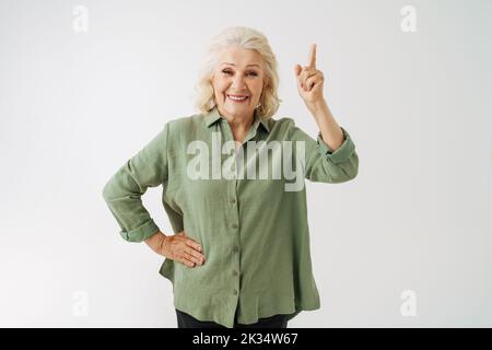 Graue ältere Frau im Hemd lächelt und zeigt den Finger isoliert auf weißem Hintergrund nach oben Stockfoto