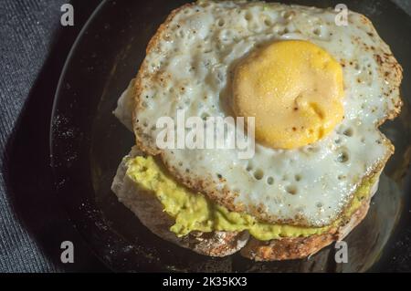 Spiegelei und auf ein Stück Brot mit Avocado, schwarzem Hintergrund und hellem Licht gelegt. Stockfoto