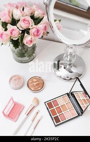Kosmetik für Make-up, runder Spiegel und Rosenblüten auf dem Tisch. Dekorative Kosmetik, Pinsel, Augenschatten mit Fundament auf Frisiertisch in Schönheit Stockfoto