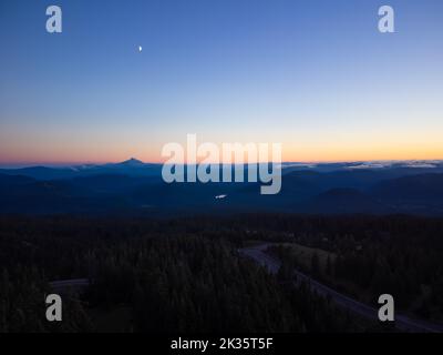 Aus der Luft schießen. Atemberaubende Nachtlandschaft. Autoweg zwischen Wald und Bergen. Der Himmel ist mit orangen Strahlen der Einstellung s gefärbt Stockfoto