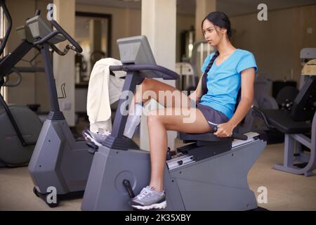 Die letzten Kilometer. Eine junge ethnische Frau, die auf einem stationären Fahrrad in der Turnhalle trainieren. Stockfoto