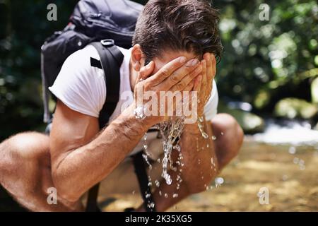Nichts erfrischender. Ein junger Mann spritzt kühles Wasser auf sein Gesicht, während er in der Nähe eines Baches wandert. Stockfoto