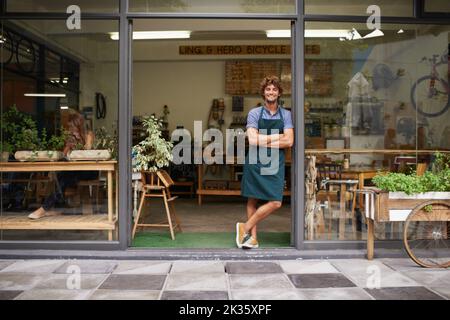 HES ist stolzer Ladenbesitzer. Ein hübscher junger Ladenbesitzer, der am Eingang seines Ladens steht. Stockfoto