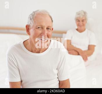 Mit einem Lächeln aufwachen. Porträt eines lächelnden älteren Mannes, der auf einem Bett mit seiner Frau im Hintergrund sitzt. Stockfoto