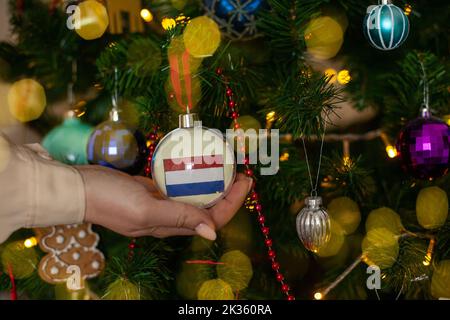 Nahaufnahme der Mädchen Hand hält einen Weihnachtsball für eine Tanne mit der Flagge der Niederlande. Neues Jahr in den Niederlanden. Weihnachtsfeiertag Grußkarte Stockfoto