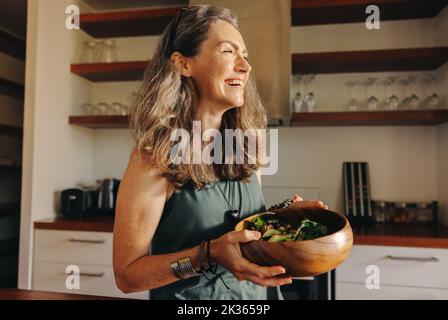 Eine alternde Frau lächelt glücklich, während sie eine buddha-Schüssel in ihrer Küche hält. Glückliche ältere Frau, die sich zu Hause ein gesundes veganes Essen serviert. Reife Frau Stockfoto