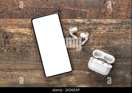 Handy mit weißem, leerem Bildschirm. Weiße kabellose bluetooth-Kopfhörer und Ladetasche und Mockup-Handy auf rustikalem Holztisch. Audio Stockfoto
