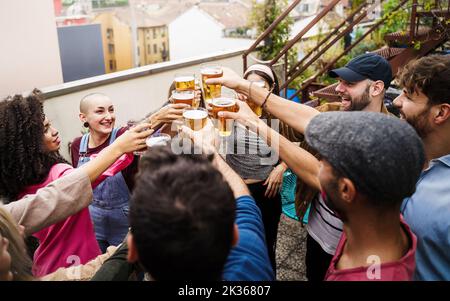 Multirassische Freunde feiern mit einem Toast klirrende Biere stehen zusammen auf der Dachparty - Gruppe von jungen Leuten, die Spaß auf der Terrasse haben - Stockfoto