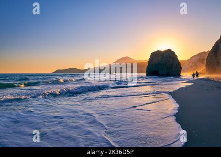 Silhouetten von zwei Erwachsenen, die bei Flut spazieren, Strand bei Sonnenuntergang, Milos, Griechenland. Ikonischer Fyriplaka einsamer Felsen gegen niedrige Sonne, farbenfrohen Himmel und Meer Stockfoto