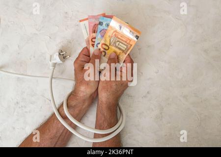 Die Hände eines Mannes, der Euro-Banknoten hält und mit einem Stromkabel mit Stecker gebunden ist, Konzept für steigende Stromkosten und Energieeinsparung CO Stockfoto