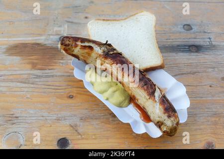 Gebratene Wurst oder deutsche Bratwurst auf einem Pappteller mit Senf und Brot auf einem alten Holztisch, typisches Outdoor-Fast-Food auf einer Ferienveranstaltung, Kopie Stockfoto