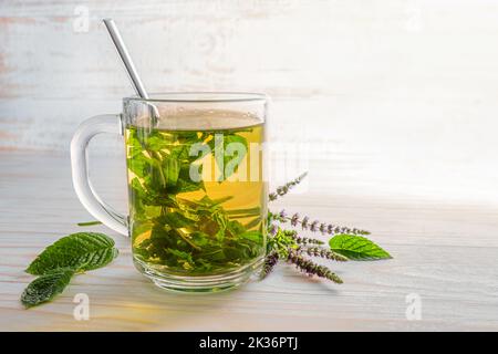 Frische Pfefferminzblätter und Blumen, die als Kräutertee in einer Glasschale auf einem hellen Holztisch gebraut werden, Kopierraum, ausgewählter Fokus, enge Schärfentiefe Stockfoto