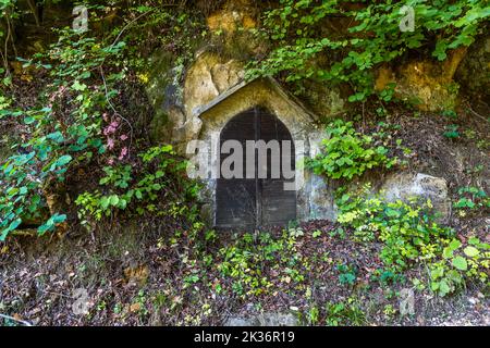 Ein Felsenkeller mit einer Holztür ist in einen Sandhügel nahe Lichtenfels, Bayern, Deutschland, gehauen. Solche in den weichen Sandstein der Region gehauenen Keller wurden traditionell in Oberfranken genutzt, um Bier zu lagern, das dort das ganze Jahr über kühl blieb. Efeu und Wildreben bedecken den Felsen um den Eingang. Stockfoto