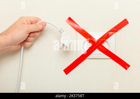 Eine mans Hand hält ein hängend weißes Kabel mit einem Stecker in der Nähe einer Steckdose, mit einem roten Band quer gebunden. Stockfoto