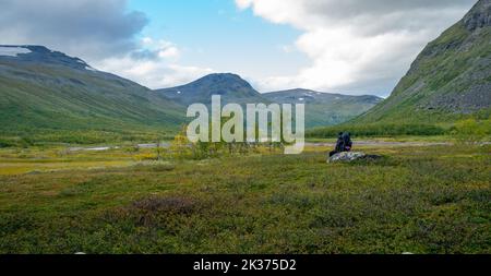 Wandererin mit schwerer Ausrüstung sitzt auf einem Felsbrocken und überblickt das weite arktische Tal Rapadalen im Sarek-Nationalpark, Lappland, Schweden. Wandertag in Stockfoto