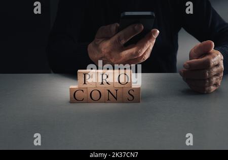 vor- und Nachteile auf Holzblöcken mit Mann, der auf blaugrauem Hintergrund auf das Mobiltelefon schaut, Konzept der Entscheidungsfindung. Stockfoto