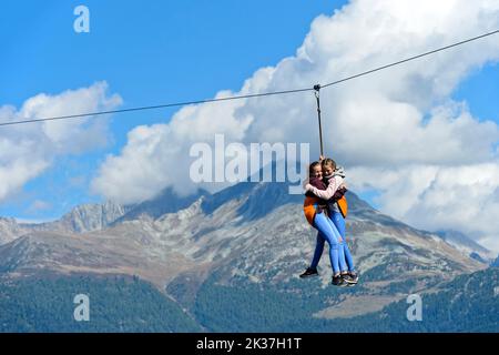 Zwei lachende Mädchen hängen am Seil einer Seilrutsche und rutschen vom Kirchturm hinunter auf den Boden, Visperterminen, Wallis, Schweiz Stockfoto