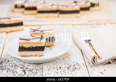 Mohn-Kuchenscheiben mit Zuckereimer, verziert mit geschmolzener Schokolade. Tortenname Berlin Kuchen. Serviert auf dem weißen Holztisch. Süßes Dessert. Stockfoto