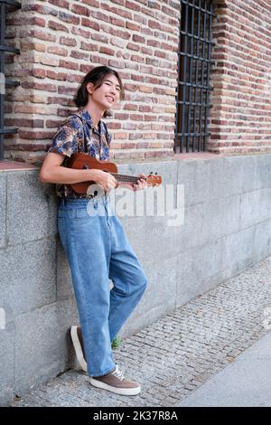 Asiatischer Kerl lächelt und spielt akustische Ukulele-Gitarre. Stockfoto