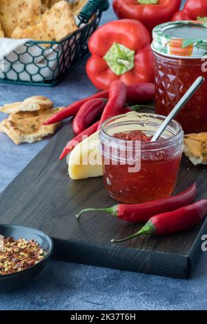 Gläser mit Paprikagelee, umgeben von Paprikaschoten, Crackerbrot und Käse. Stockfoto