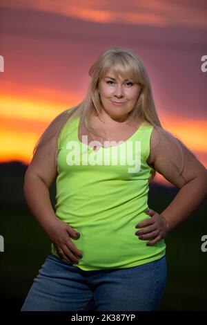 Eine dicke Frau mittleren Alters posiert in Jeans und einem hellen T-Shirt vor dem Hintergrund des Abendhimmels, übergewichtig xxl. Ein volles Mädchen genießt das Leben. Stockfoto