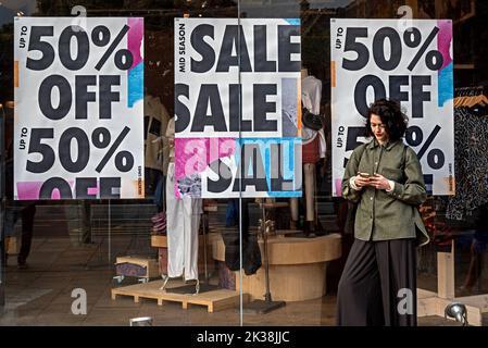 Junge Frau, die ihr Telefon mit 50 % Rabatt-Schilder auf dem Schaufenster hinter ihr anschaut. Edinburgh, Schottland, Großbritannien. Stockfoto