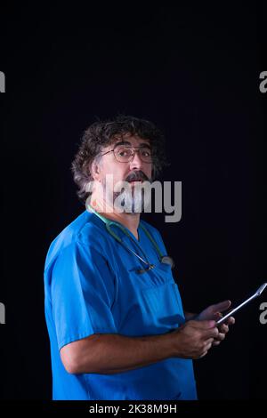 Arzt mit einem Bart und einer Brille in einem blauen OP-Kittel, der den Patienten anschaut, während er einen Tisch mit dem medizinischen Datenprofi hält Stockfoto