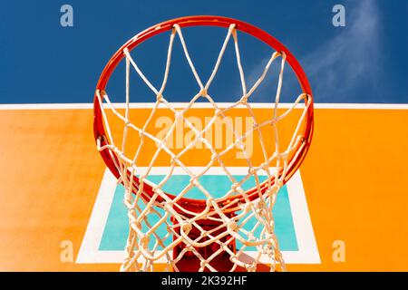 Low-Angle-Ansicht des farbenfrohen orangen Basketball-Backboards mit rotem Reifen und einem Netz gegen den blauen Himmel. Stockfoto