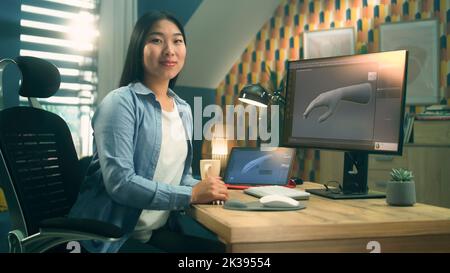 Weibliche asiatische 3D Designerin sitzt am Tisch in der Nähe des modernen Computers und digitalen Tablets, arbeitet an 3D Projekt aus der Ferne von zu Hause aus und schaut auf die Kamera. Stockfoto