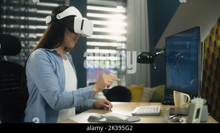 Asiatische Frau, die im Home Office am Tisch sitzt und mit einem VR-Headset an einem Modellierungsprojekt von 3D am PC arbeitet. Virtuelle Realität. Metaverse Stockfoto