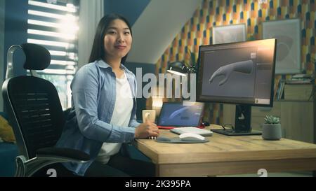 Weibliche asiatische 3D Designerin sitzt am Tisch in der Nähe des modernen Computers und digitalen Tablets, arbeitet an 3D Projekt aus der Ferne von zu Hause aus und schaut auf die Kamera. Stockfoto