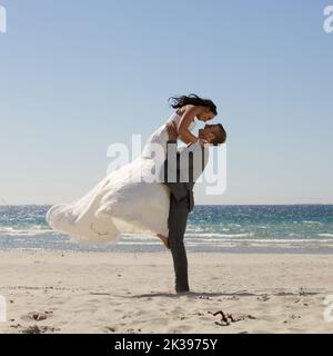 Der Bräutigam hebt die Braut an einem Strand mit Wellen im Hintergrund Stockfoto