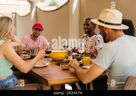 Ein schwarzer Mann und ein kaukasischer Mann mit roter Mütze hören lächelnd zu, wie sie mit Freunden das Mittagessen feiern. Stockfoto