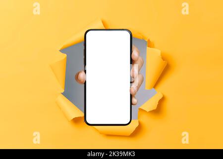 Männliche Hand zeigt das Smartphone mit weißem Bildschirm durch Loch in gerissenem orangefarbenem Papierhintergrund. Mockup für App, Werbung oder Präsentation. Copy-Space-Konzept Stockfoto