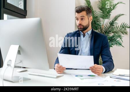Schockierter kaukasischer Geschäftsmann, Firmenchef, Finanzmanager, in einem eleganten Anzug gekleidet, sitzt im Büro am Arbeitsplatz, hält Finanzdokumente in der Hand, sieht überrascht auf den Computerbildschirm Stockfoto