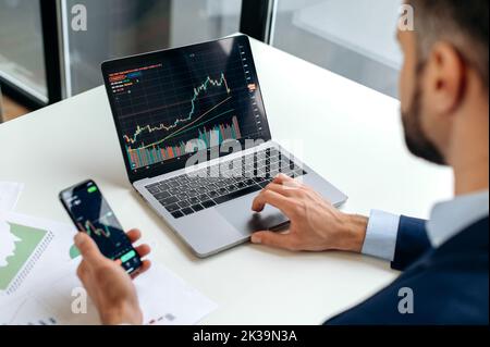 Kaukasischer Krypto-Trader-Investor, der Mobiltelefon und Laptop für die Kryptowährung-Finanzmarktanalyse verwendet, Kryptowährung kauft oder verkauft.Nahaufnahme des Laptops und Smartphones mit Aktiendiagrammen Stockfoto