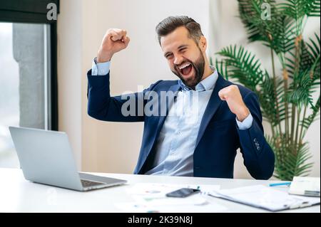 Victory-Konzept. Erfolgreicher fröhlicher glücklicher kaukasischer bärtiger Geschäftsmann, ceo Firma, Manager, sitzt am Schreibtisch im Büro, gestikuliert mit Fäusten, feiern Erfolg, gewinnen, großer Gewinn, Stellenangebot Stockfoto