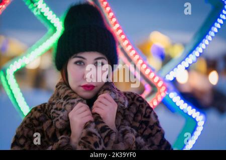 Porträt einer jungen Frau in schwarzem Hut und Pelz Mantel mit Leopardenmuster steht auf dem Hintergrund der Weihnachtsbeleuchtung Dekorationen und Blick auf die Kamera Stockfoto