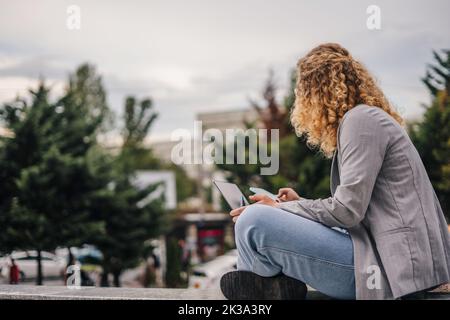 Seitenansicht einer jungen College-Studentin in grauer Jacke, die draußen an der Universität mit ihrem Laptop studiert. Freier Speicherplatz für Text. Entfernt Stockfoto