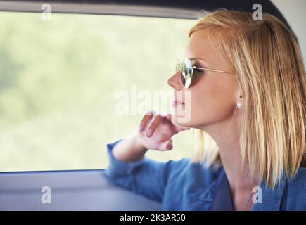 Viel Zeit zum Nachdenken auf offener Straße. Eine junge blonde Frau, die aus dem Fenster schaut, während sie in einem Auto sitzt. Stockfoto
