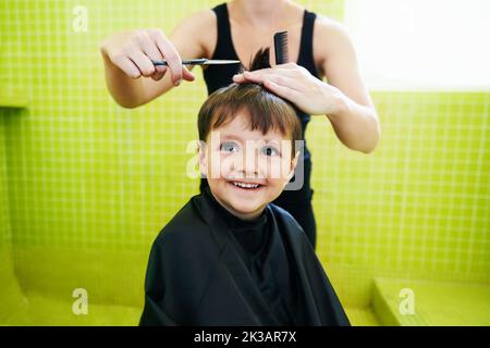 Glücklich mit seinem neuen Haarschnitt. Ein kleiner Junge lächelt, als er sich die Haare schneiden lässt. Stockfoto