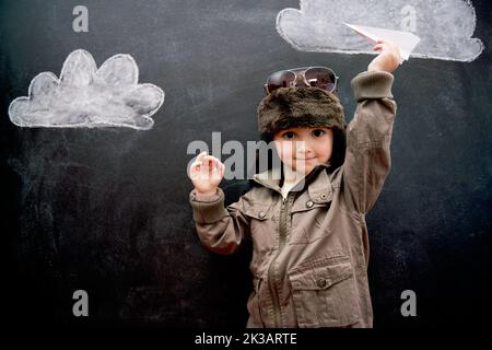 Dieses Kind geht auf die Plätze. Ein kleiner Junge spielt mit einem Flugzeug vor Wolken auf einer Tafel gezeichnet. Stockfoto