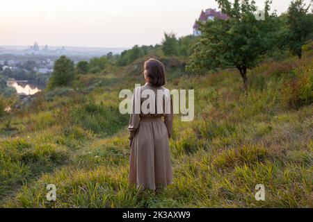 Schöne junge lächelnde Mädchen in einem langen braunen Kleid steht entlang des Rasens. Glückliche Frau geht bei Sonnenuntergang auf einem Hügel mit Blick auf den Fluss. Konzept des havi Stockfoto