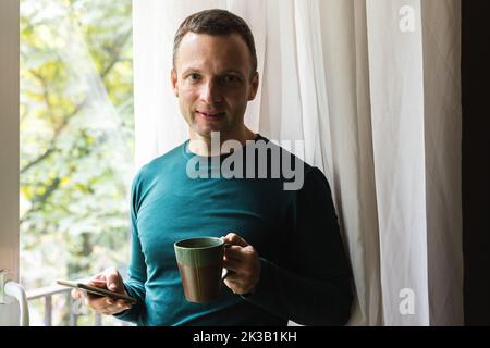 Casual Home Portrait eines leicht lächelnden jungen erwachsenen kaukasischen Mannes mit einer Tasse Kaffee und einem Smartphone in den Händen Stockfoto