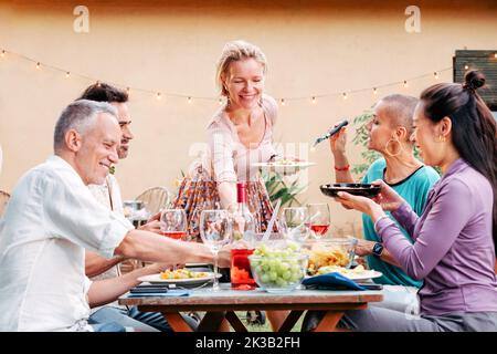 Eine Erwachsene blonde Frau lächelte und servierte Teller mit Salat auf dem Tisch, verschiedene Freunde saßen herum und hatten Spaß. Lifestyle-Konzept. Stockfoto