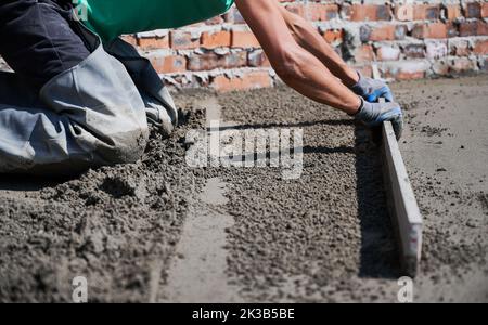 Nahaufnahme eines Bauherrn, der auf der Baustelle Estrich-Schienen auf den mit Sand-Zement-Gemisch bedeckten Boden legte. Männlicher Arbeiter nivellieren Fläche mit gerader Kante während Estrichboden im neuen Gebäude. Stockfoto