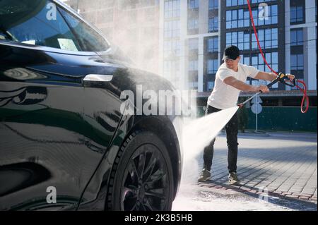 Guy in Cap Reinigung seines schwarzen Autos mit Hochdruckwasser im Service. Waschen von Auto mit fließendem Wasser auf dem Hintergrund des Wohngebäudes. Stockfoto