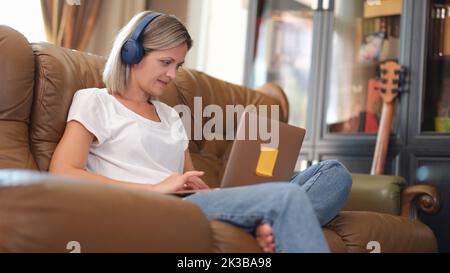 Die attraktive junge Frau sitzt auf der Couch und nutzt Laptop und Kopfhörer Stockfoto