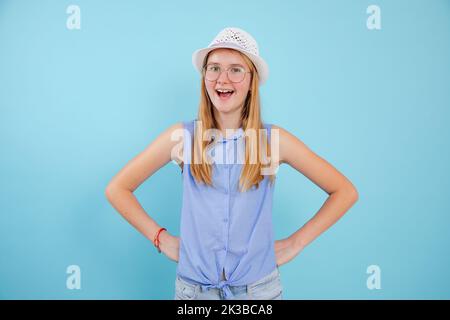 Porträt eines Teenagers mit offenem Mund, weißem Sommerhut, Brille und Händen auf blauem Hintergrund. Stockfoto