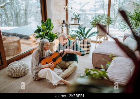 Seniorenpaar spielt auf der Gitarre, sitzt im gemütlichen Wohnzimmer und genießt den Herbsttag. Stockfoto